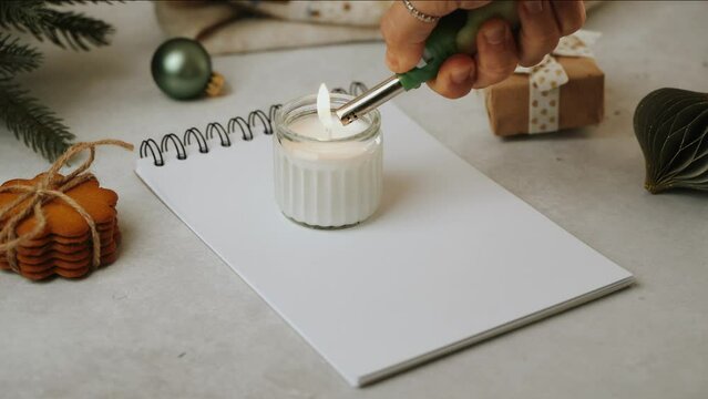 Christmas goals. Close up woman's hands light a candle. Blank notepad for new year resolution . Still life of white working table background with candle and cozy Christmas decoration. 