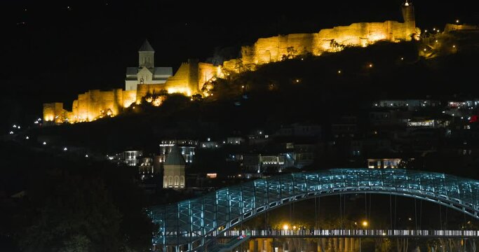 Tbilisi, Georgia. Night Scenic View Of Impregnable Narikala Fortress And Bridge Of Peace Over Kura Mtkvari River In Evening Illuminations. Bridge Of Peace At Evening Night Street Illumination.