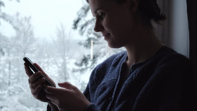 Close up profile of attractive young female sitting on comfy windowsill and holding mobile phone enjoying beautiful winter nature. Smartphone application market, podcast listening. Relaxation concept
