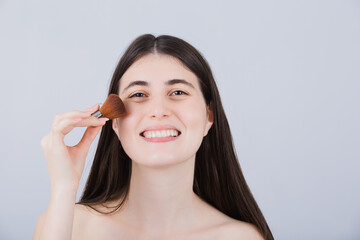Smiling Young woman applying cosmetics with makeup brush. Happy young woman using cosmetic brush applying face powder looking to camera.