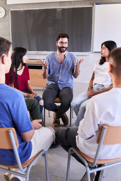 Vertical Of Diverse Group Of High School Students Sitting On Chairs In Circle And Interacting During A Lesson, Their Caucasian Male Teacher With Them And Talking Education Concept. High Quality Photo