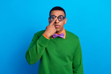 Portrait photo of funny childish guy wear green jumper shirt purple bowtie fix his eyeglasses interested looking you isolated on blue color background