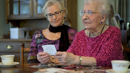 Low angle extreme closeup of senior elderly happy smiling woman looking at old photos and remembering memories with daughter at the dining room table.