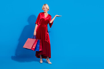 Full length photo of dreamy sweet retired lady dressed shiny red clothes holding shoppers arm empty space isolated blue color background