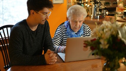 Teen grandson teaching elderly senior grandmother how to use a laptop computer in a dining room.