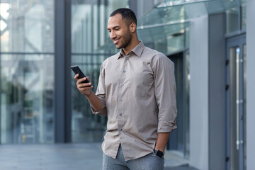 A young handsome Hispanic, African-American man is standing on the street near an office center. He holds the phone in his hand, uses it, dials, smiles.