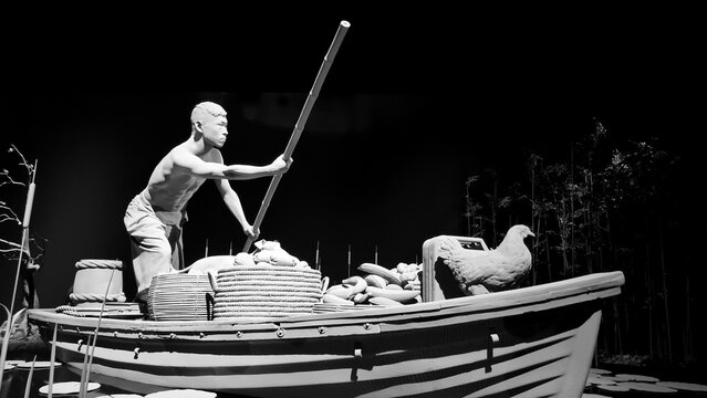 Grayscale of a boatman on a boat full of items on the water by Hans Op De Beeck