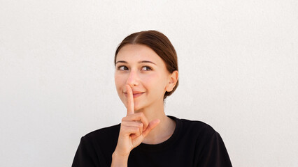 Portrait of positive young woman making secret gesture over white background. Caucasian woman wearing black T-shirt looking away with finger on lips. Secret and silence concept