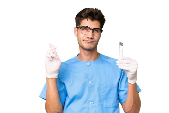 Young dentist man holding tools over isolated background with fingers crossing and wishing the best