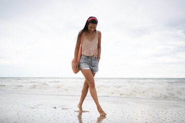 Chica morena en pantalón corto y camiseta de tirantes caminando sobre la arena mojada de la playa tras el paso de una ola