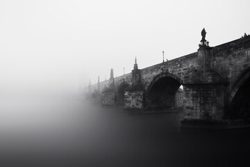 Charles bridge from below in the fog in the early morning in Prague with statues and lanterns on the bridge. Black and White. Czech Republic.