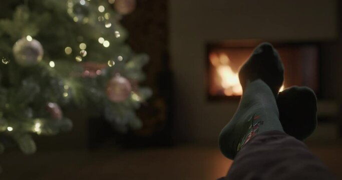 A man sits by a fireplace and a Christmas tree, first-person view. Feet in festive socks are visible in the frame