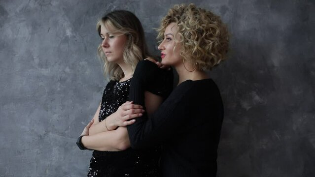 Thoughtful women stand half turn and hug each other isolated on gray wall background. Place for text. Old friends talking. Beautiful female wear for holidays. Celebration, party, festive look