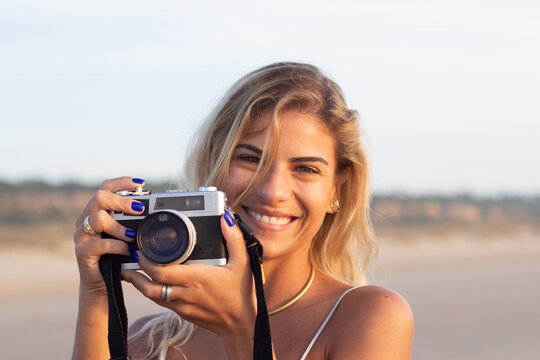 Portrait of beaming woman with old-fashioned camera. Female model with fair hair and in casual clothes holding camera on beach. Hobby, travelling concept