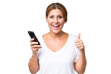 Fototapeta premium Middle-aged caucasian woman over isolated background using mobile phone while doing thumbs up