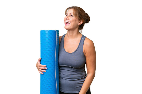 Middle-aged sport woman going to yoga classes while holding a mat over isolated background laughing in lateral position
