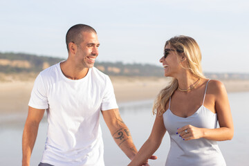 Smiling wife and husband during honeymoon. Man with shaved head and woman in casual clothes looking at each other, walking on beach. Love, vacation, family concept