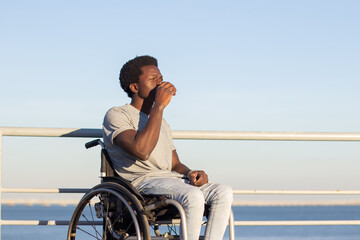 Young man with disability having takeaway coffee outside. Short-haired Black guy in wheelchair enjoying espresso against sea background on sunny summer day. Low angle. Disability, lifestyle concept.