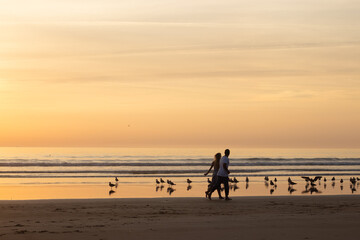 Fototapeta na wymiar Warm couple walking on beach at sunset. Man and woman in casual clothes running along water at dusk. Love, family, nature concept