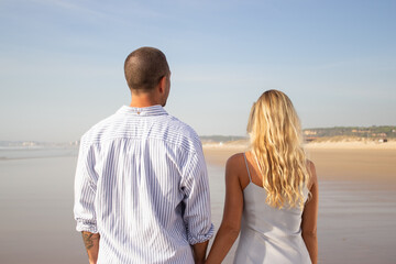 Back view of couple walking on beach. Caucasian man with shaved head and woman in casual dress holding hands. Love, vacation, affection concept