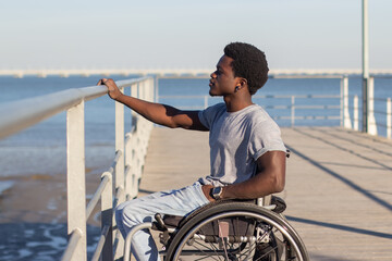 Fototapeta premium Young Black man in wheelchair looking at seascape. Side view of confident stylish guy with paraplegia laying his hand on railing, enjoying sea view. Medium shot. Disability, attitude concept.