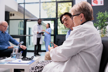 Elderly medic measuring glucose and insulin level with glucometer while doing sugar test for asian patient with diabetes. Young adult having medical appointment in hospital waiting area.