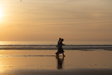 Naklejka premium Romantic couple walking on beach at sunset. Man and woman in casual clothes strolling along water at dusk. Love, family, nature concept