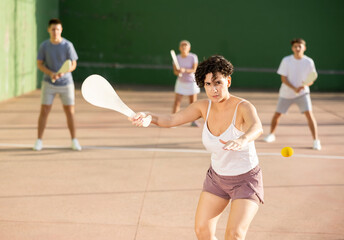 Hispanic woman pelota player hitting ball with wooden racket during training game on outdoor Basque pelota fronton.