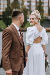 A stylish groom in a brown suit and a beautiful smiling model blonde bride are walking in the city,...