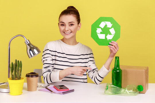 Delighted woman office manager holding recycling sign and pointing at plastic, glass bottles and cardboard package, thinking green. Indoor studio studio shot isolated on yellow background.