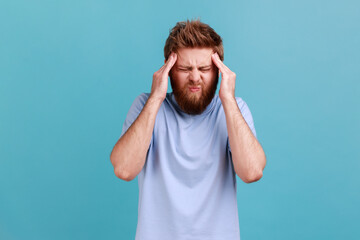 Fototapeta na wymiar Portrait of unhealthy bearded man massaging temples feeling headache, suffering migraine or high blood pressure, trying to concentrate. Indoor studio shot isolated on blue background.