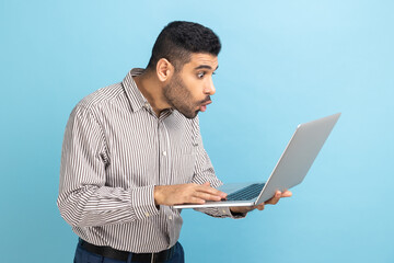 Portrait of shocked businessman with beard looking at laptop display with opened mouth and big eyes, system error, wearing striped shirt. Indoor studio shot isolated on blue background.
