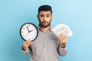 Portrait of upset sad businessman holding big fan of dollar banknotes and wall clock, time is...