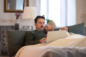 Gay lying in bed and sleeping on his lover shoulder in morning. Focused homosexual man working on laptop while lying under blanket in bed with partner. LGBT, morning routine, love concept