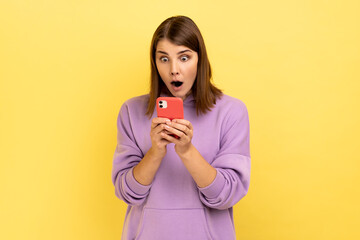 Portrait of amazed excited woman reading message on smartphone, using mobile device for communication, surprised with app, wearing purple hoodie. Indoor studio shot isolated on yellow background.