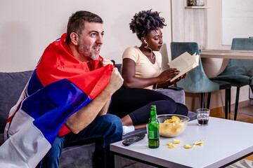 The husband is watching a football match on TV, while his wife is bored. The woman is trying to read a book with earbuds in her ears. World cup. Marriage agreements.