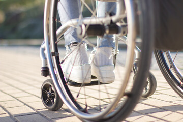 Person of unrecognizable gender and age sitting in wheelchair outdoors on sunny day. Close-up shot of thin legs in white sneakers and light blue jeans. Disability concept. - obrazy, fototapety, plakaty