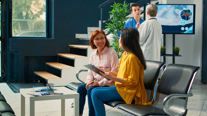 Asian patients talking about medical report papers to receive insurance support, sitting in waiting room at facility. Women preparing to attend checkup visit, filling in medication form.