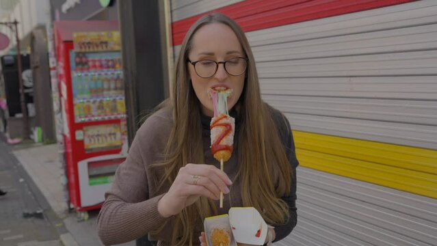 This video shows a traveling caucasian woman eating a rainbow colored cheese corn dog on the streets of japan.