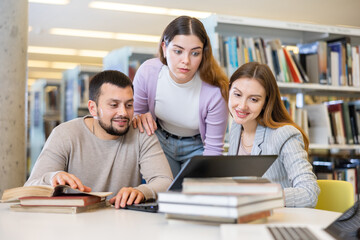Group of positive friendly international university students working on joint research in library, looking for information in books and websites using laptop