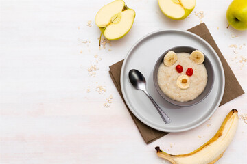 Funny cute kids childrens baby's healthy breakfast lunch oatmeal porridge in bowl look like bear face decorated with apple, banana, dried berry fruits. dessert food art on white wooden table