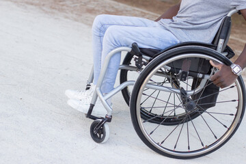 Tilted closeup shot of young Black man in wheelchair outside. African American guy in white sneakers and blue jeans enjoying ride in city park on warm day. Disability, motivation concept.
