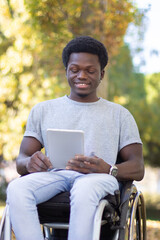 Fototapeta premium Young Black man in wheelchair spending time outdoors in park, reading digital tablet, smiling and having fun on sunny autumn day. Full shot. Modern technologies, leisure time, disability concept.
