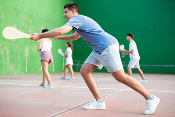 Portrait of emotional determined young guy playing pelota at open-air fronton in summer, swinging...