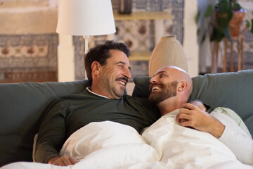 Happy gays laughing while lying in bed in morning. Medium shot of handsome Caucasian men looking at each other and smiling, having fun together after waking up on weekend. LGBT, sleep cycle concept