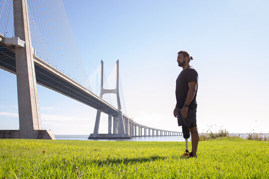 Side view of bearded man with amputated leg enjoying summer day. Sporty man standing on grassy field near sea resting after running looking far. Healthy lifestyle of people with disability concept
