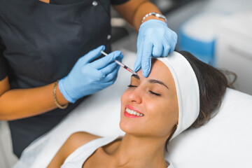 Eyebrow filler injection concept. Closeup indoor portrait of smiling caucasian woman with her head placed on SPA bed during collagen injection performed by professional expert in protective gloves