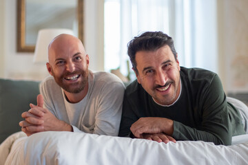 Obraz premium Happy gays lying on bed and smiling at camera. Front view of cheerful bearded homosexual men in pajamas spending morning together, posing for camera. LGBT, sleep cycle, togetherness concept