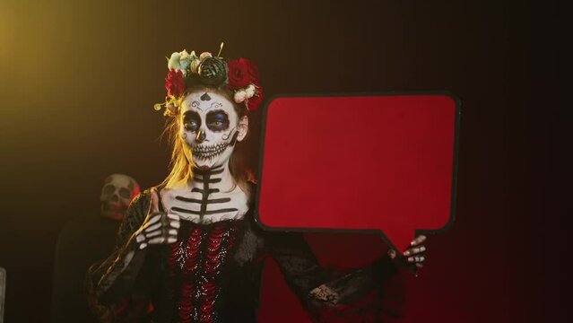 Cavalera catrina showing isolated speech bubble cardboard, wearing santa muerte costume and skull make up. Person using blank copyspace template on carton board, holy goddess of death. Handheld shot.
