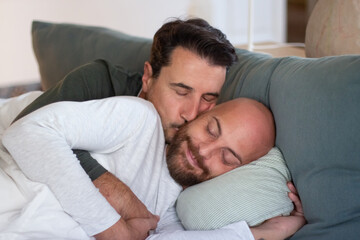 Gay lying in bed with partner in morning and kissing him. Medium shot of cheerful bearded man lying with closed eyes and hugging his lover after waking up. LGBT, love, sleep cycle concept